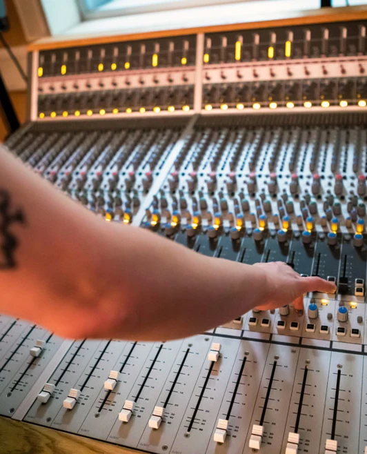 A close-up of a student's arm with a ghost tattoo reaching for controls on a large mixing desk, with glowing indicator lights. A close-up of a student's arm with a ghost tattoo reaching for controls on a large mixing desk, with glowing indicator lights.
